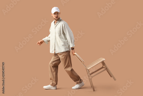Obraz na plátne Stylish young man in eyeglasses with cap carrying chair on beige background
