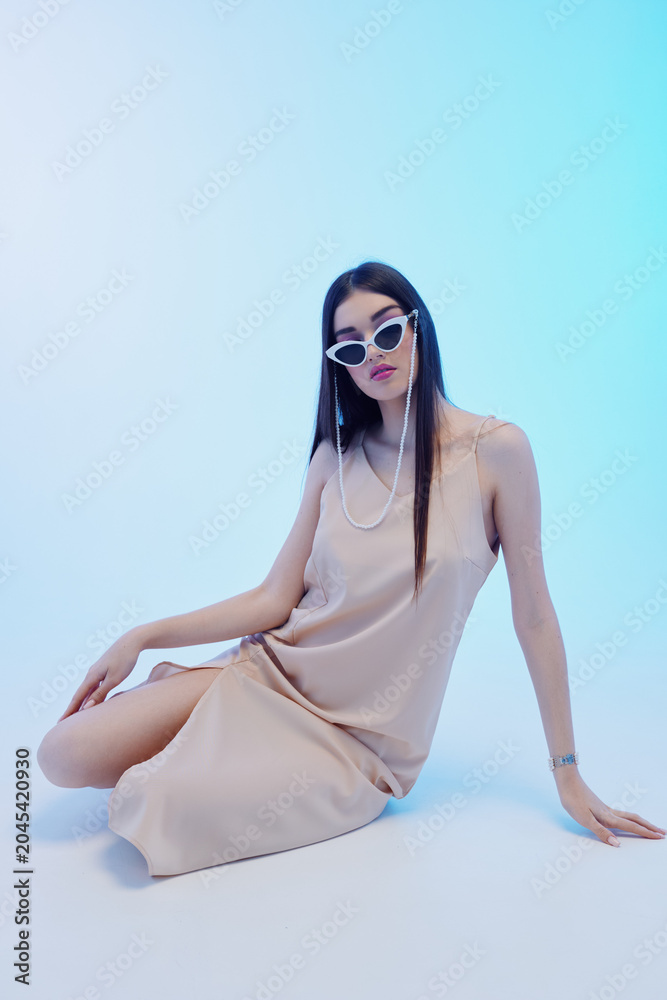 Naklejka premium Young woman in a beige dress and white sunglasses poses seated on a light blue background. Concept of beauty, fashion, and modern style with elegant minimalism.