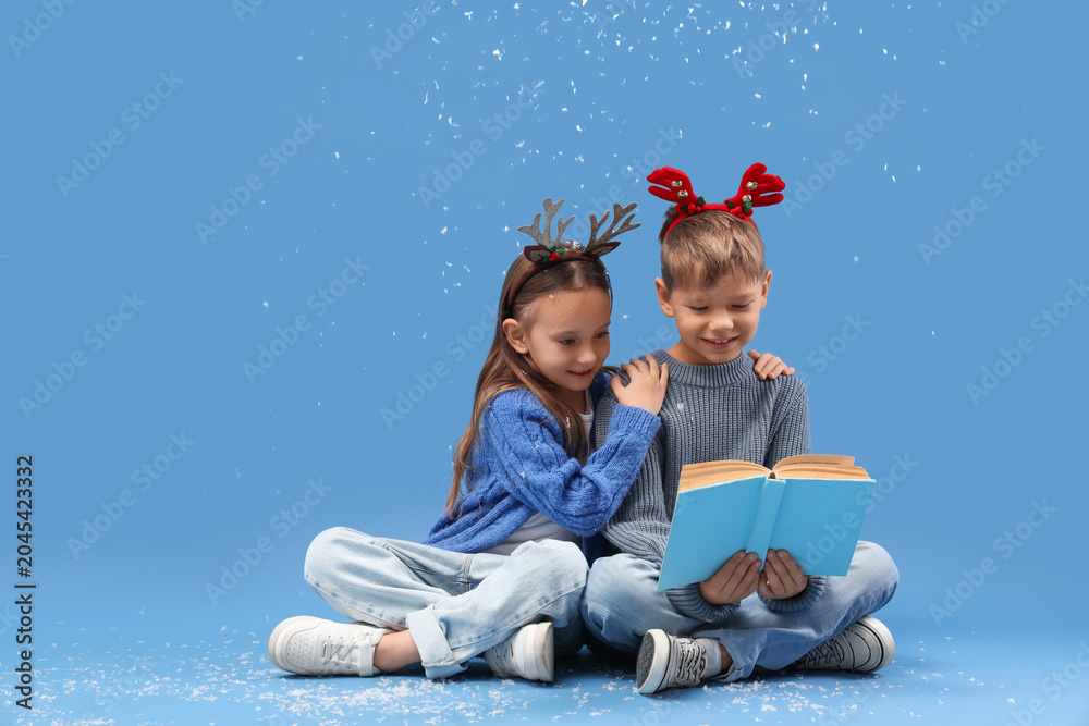 Naklejka premium Cute little children in Christmas reindeer horns with snow reading book on blue background