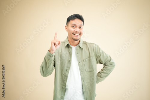 Young Asian man holding up one index finger with a eureka expression, hand on hip, smiling brightly suggesting an idea in casual studio portrait