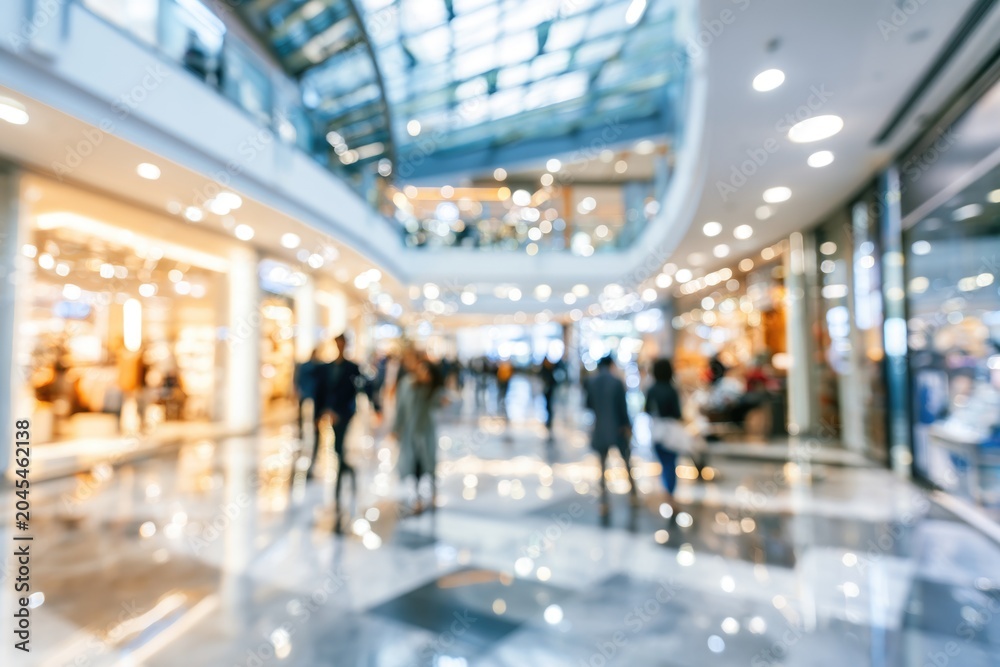 Fototapeta premium A blurred image of a bright shopping mall interior with people walking and illuminated store windows.