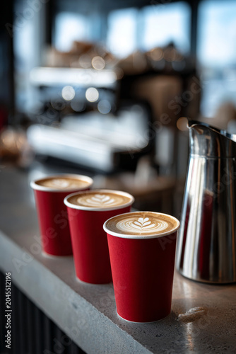 Close-up of three identical red paper takeaway coffee cups filled with latte art, arranged in a triangular formation on a clean cafe counter with a coffee machine in the background