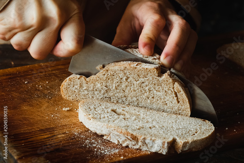 Hands slicing freshly baked loaf of brown bread on cutting board