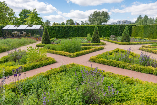 A formal parterre garden shows boxwood hedges, conical topiary, herb beds, lavender, and a greenhouse. Brick paths, vegetable rows, and clipped greenery create an orderly ornamental layout.