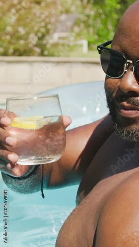 Vertical video: Cam filming shirtless man in shades raising glass, lemon in backyard pool toasting
