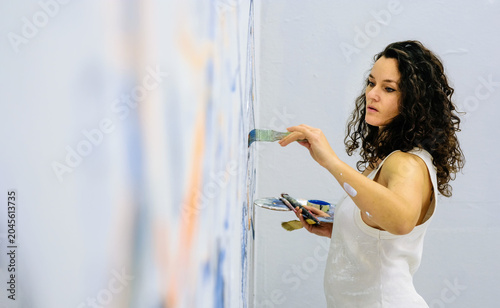 Female artist painting a large wall mural in a bright studio while holding brushes and palette. Creative process, concentration and contemporary visual art in progress