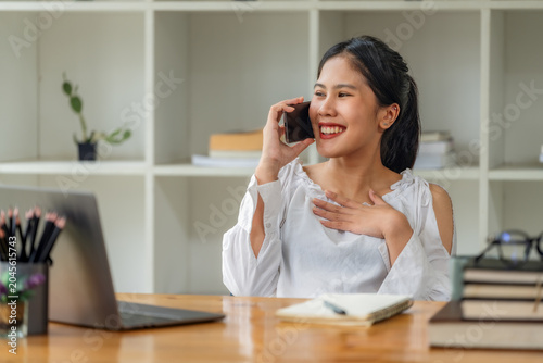 Happy young Asian businesswoman talking on smartphone while working with laptop at office desk.