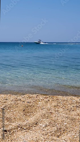 Sunny Beach With Clear Blue Water, Pebble Shore, And Speedboat On The Horizon