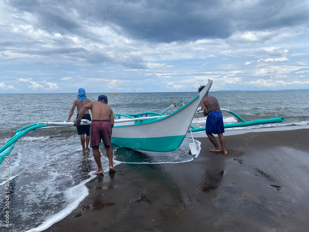 Obraz premium Fishermen are launching a small fishing boat from the shore amid rough sea waves, with a typhoon approaching the small fishing village.