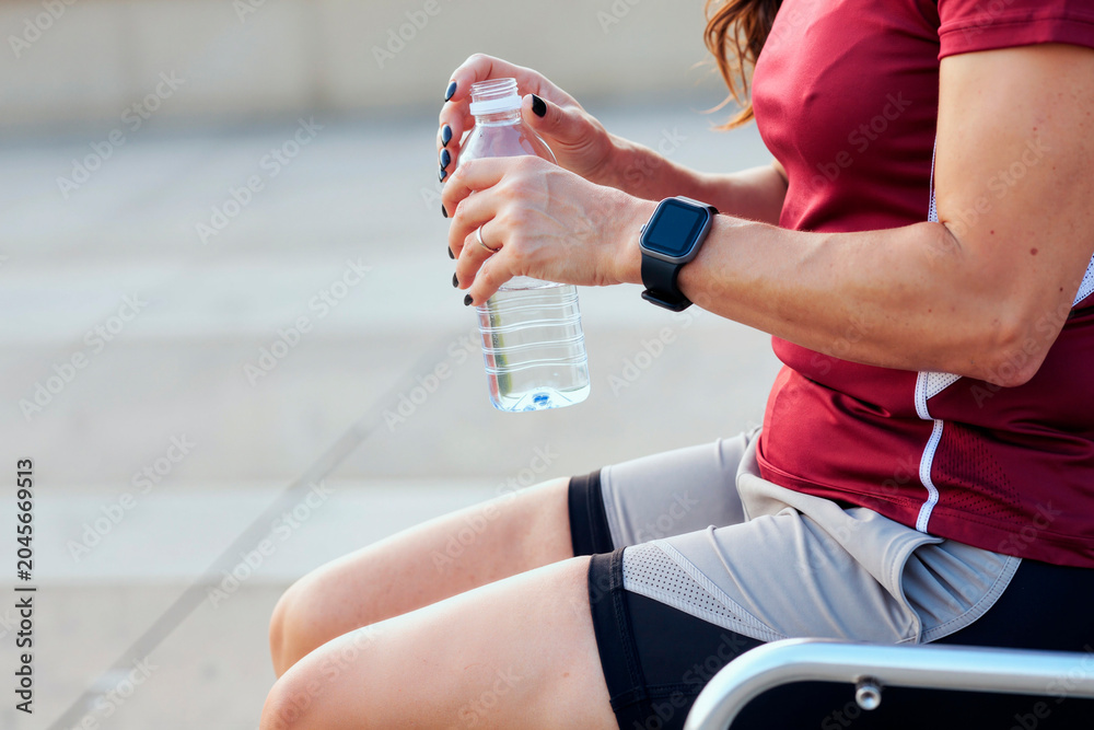 Naklejka premium Caucasian middle aged woman resting after workout, opening water bottle and wearing smartwatch, showing hydration and fitness tracking. Ideal for wellness, active lifestyle marketing