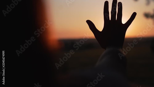 Person raises hand toward sunset creating silhouette at horizon. Human reaches palm against golden sun at dusk. Hand blocks sunlight. Person shows gesture at sunset. Silhouette reaches toward sun.