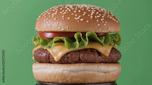 Freshly made beef burger with melted cheese, lettuce, and tomato rotating. Classic fast food meal presented on a vibrant green background