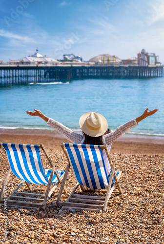 Canvastavla A happy woman sitting in a deck chair looks at the beach and pier of Brighton, E