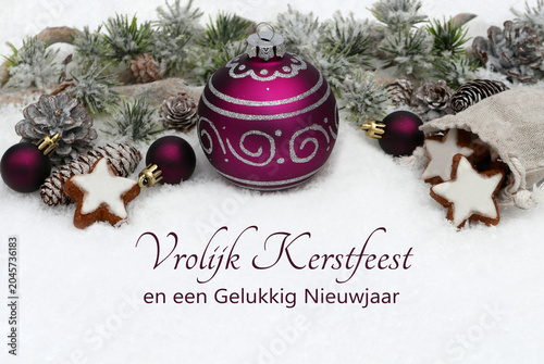 Photos Paarse kerstbal omgeven door dennenappels, kaneelsterren en kleine paarse bolletjes op sneeuw met de Nederlandse kerstgroet Vrolijk Kerstfeest en een Gelukkig Nieuwjaar