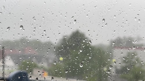 Close-up of many clear raindrops on a window pane during a heavy downpour, with a blurry background of green trees and buildings under a grey, overcast sky.
