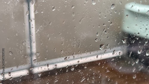 Close-up shot of raindrops collecting on a window pane during a heavy storm, with a blurry background of trees and buildings under a grey, overcast sky.