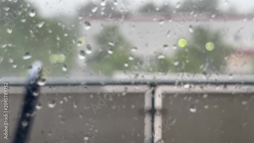 Close-up shot of raindrops collecting on a window pane during a heavy storm, with a blurry background of trees and buildings under a grey, overcast sky.
