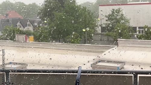 View from a window of heavy rain falling on an apartment balcony with a blue plastic chair, looking out towards green trees and a building under a overcast sky.
