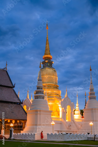 The golden pagoda of Wat Suan Dok temple illuminated at twilight on a rainy season night with a Buddhist monk standing by the wall in Chiang Mai, Thailand
