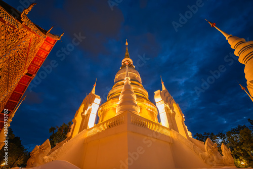 The golden pagoda of Wat Suan Dok temple illuminated at twilight on a rainy season night with a Buddhist monk standing by the wall in Chiang Mai, Thailand