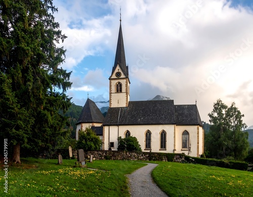 A stone church with a tall steeple and dark roof stands on a grassy knoll, surrounded by trees and a path