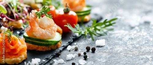 Obraz  z motywem Close-up view of buffet appetizers and salad on a black slate surface. Appetizers include shrimp canapes and cucumber-tart. Ideal for catering promotions