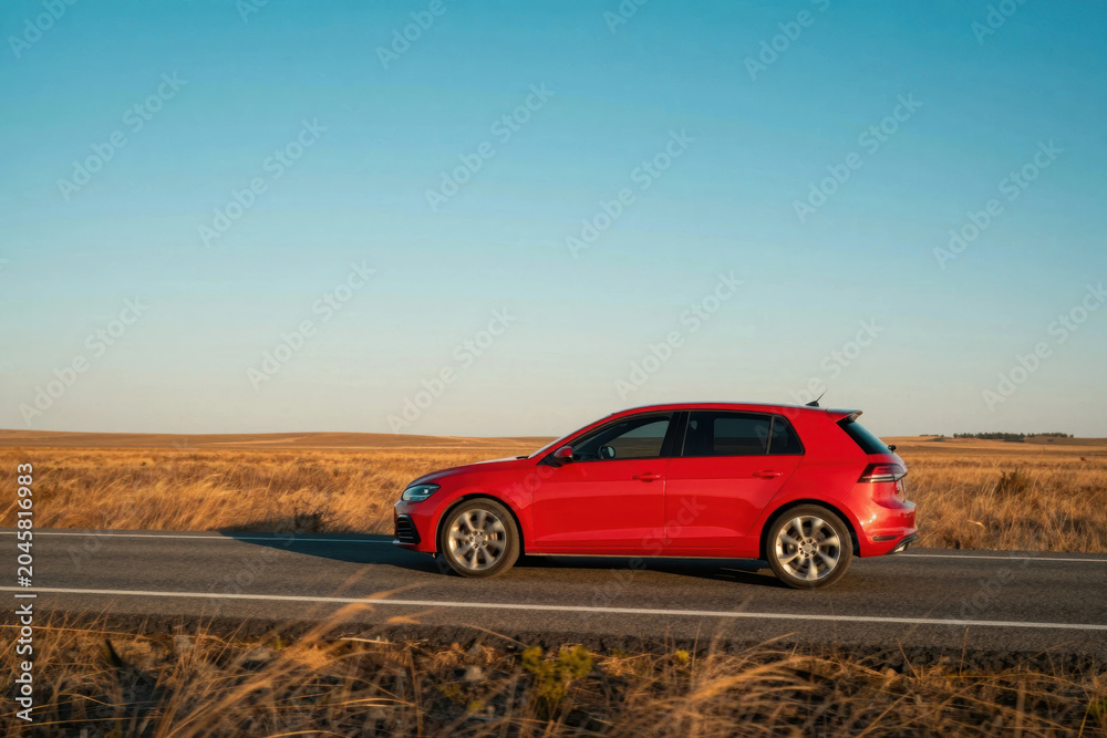 Fototapeta premium Red car drives on empty road during sunset in wide open space