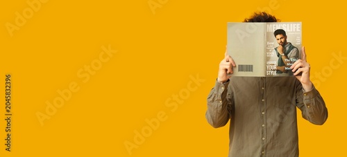 Young man covering face with lifestyle magazine while standing against bright orange background, anonymous reader holding open publication, fashion and media concept with copy space