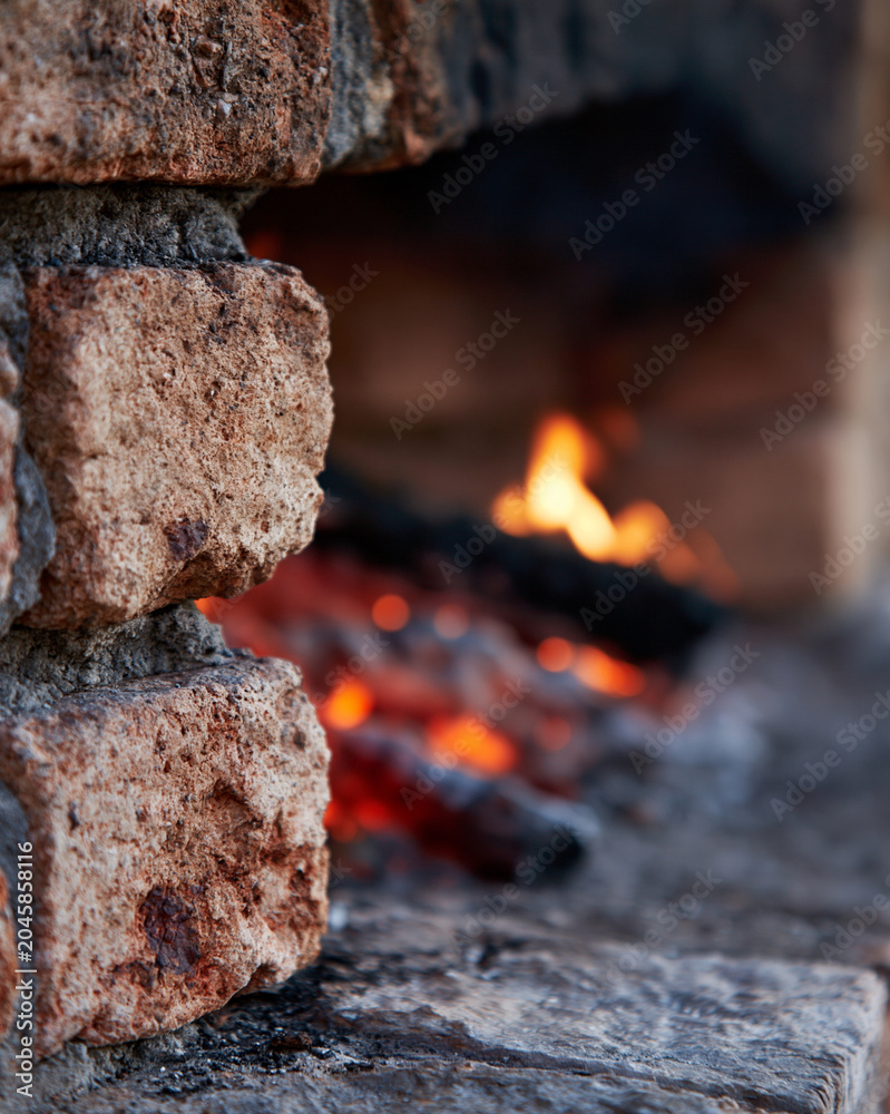 Fototapeta premium Warm fire glows within rustic stone fireplace. Embers flicker softly, casting amber light. Brick edges show weathered, textured surfaces. Cozy, inviting atmosphere radiates from hearth