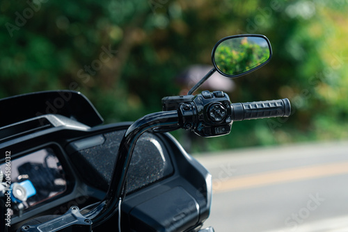 Close up of motorcycle handlebar with rearview mirror, showcasing grip and control buttons against blurred outdoor background