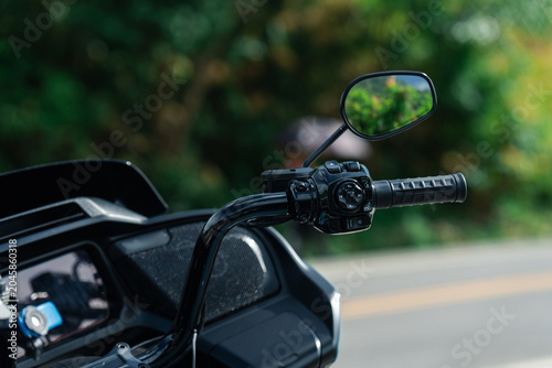 Close up of motorcycle handlebar with mirror, showcasing sleek design and black finish. background is blurred greenery