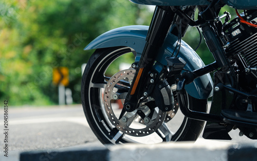 Close up of motorcycle front wheel with disc brake, parked on road with blurred greenery in background
