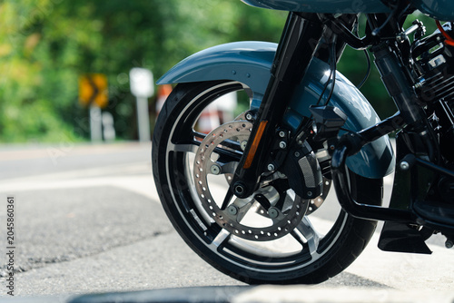 Close up of motorcycle front wheel with shiny disc brake, parked on road with blurred greenery in background
