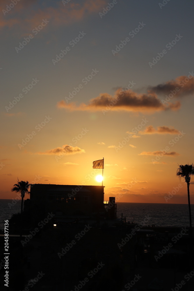 Naklejka premium Caesarea, Israel - JUNE 2, 2026: A flag waves atop a silhouetted building against a vivid orange sunset over the ocean, with palm trees framing the scene.