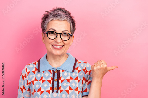 Obraz na plátně Smiling mature woman with glasses pointing to the side against a pink background
