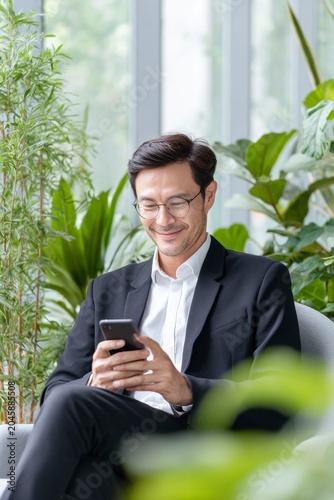 a man in a suit and glasses sits comfortably in a chair, smiling at the screen of his smartphone