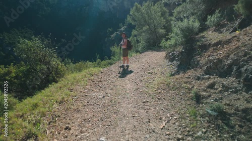 Woman hiking along sunlit mountain trail overlooking a forested canyon with backpack and poles