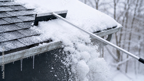 Clearing snow from a house roof with a roof rake. Winter home maintenance to prevent ice dams. Close up of snow sliding off shingles over a gutter with icicles