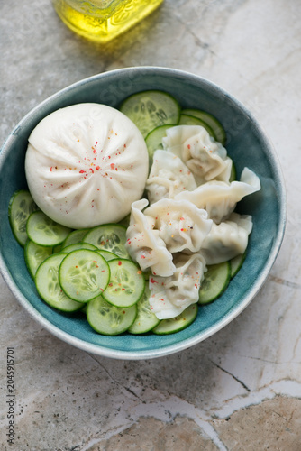 Steamed baozi with cucumber and wontons in a green bowl, vertical shot on a grey granite background, high angle view