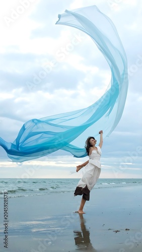 Woman dances on beach, flowing fabric billowing above her against cloudy sky, sand mirroring the light