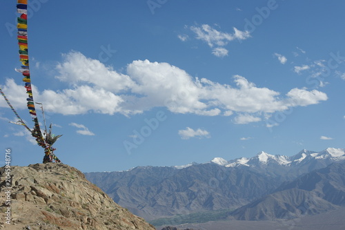 Colorful Tibetan Buddhist prayer flags fluttering on a rocky hill against majestic snow-capped mountain ranges under a blue sky in Ladakh, Himalayas.