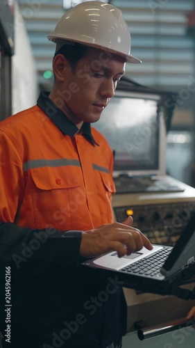 Industrial Technician at Work: A focused industrial technician, wearing a hard hat and high-visibility vest, operates a laptop in a manufacturing facility, overseeing complex machinery.