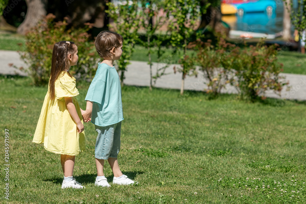 Fototapeta premium Two children, girl in yellow dress and a boy, stand hand in hand on a grassy area in a park, surrounded by trees and colorful water vehicles in the background. Happy childhood concept. Summer vacation