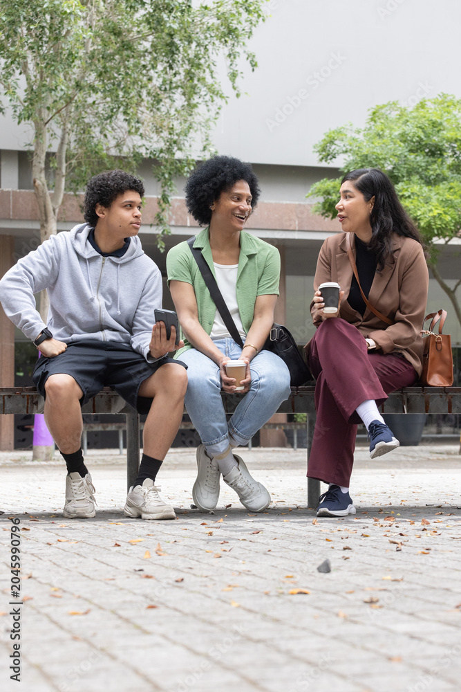 Fototapeta premium Diverse classmates chatting, sipping coffee with smartphone on wooden bench in campus plaza