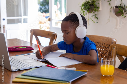 African American child with headphones sitting at dining table writing with orange pencil by laptop