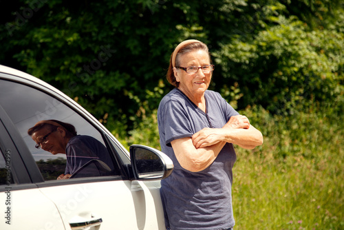 Old woman driving a car in the countryside, road trip concept.  Portrait of an old woman driving and near a car, on a walk outside the city.