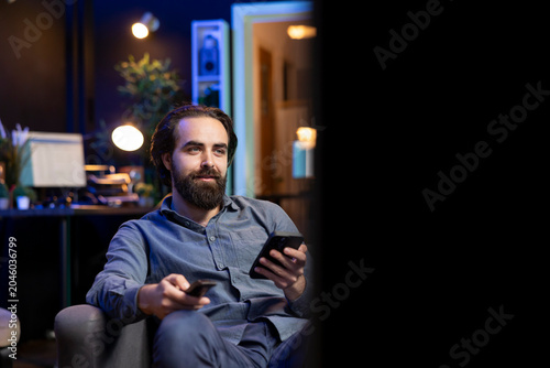 Foto Smiling person on couch binging series on streaming service, browsing using remote and texting on mobile phone