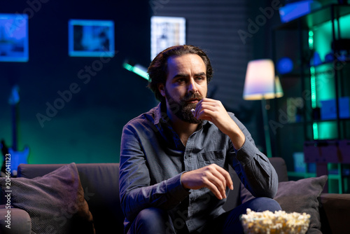 Fotografía Puzzled man eating popcorn while watching confusing movie on streaming service, enjoying time off from work