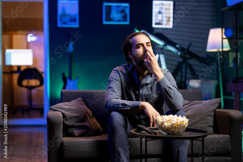 Obraz na plátně Man relaxing at home on couch, stuffing mouth with popcorn while watching movies on TV in apartment