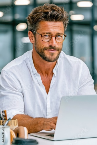Confident professional man working on laptop at modern office desk in casual white shirt